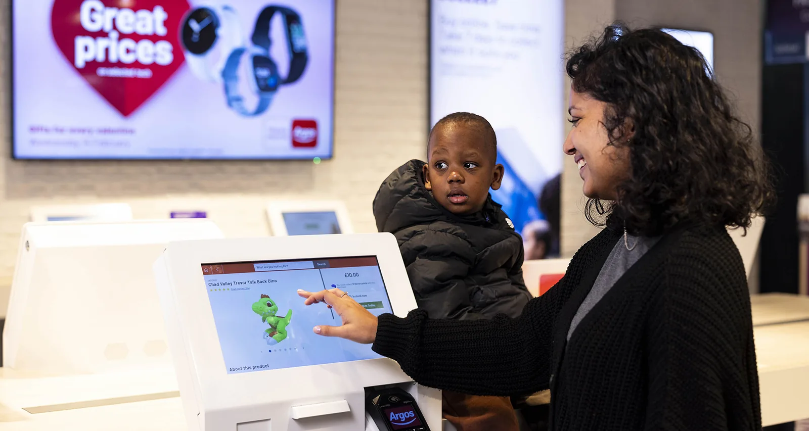 A woman holding a child reaches for a self-service ordering kiosk inside an Argos store