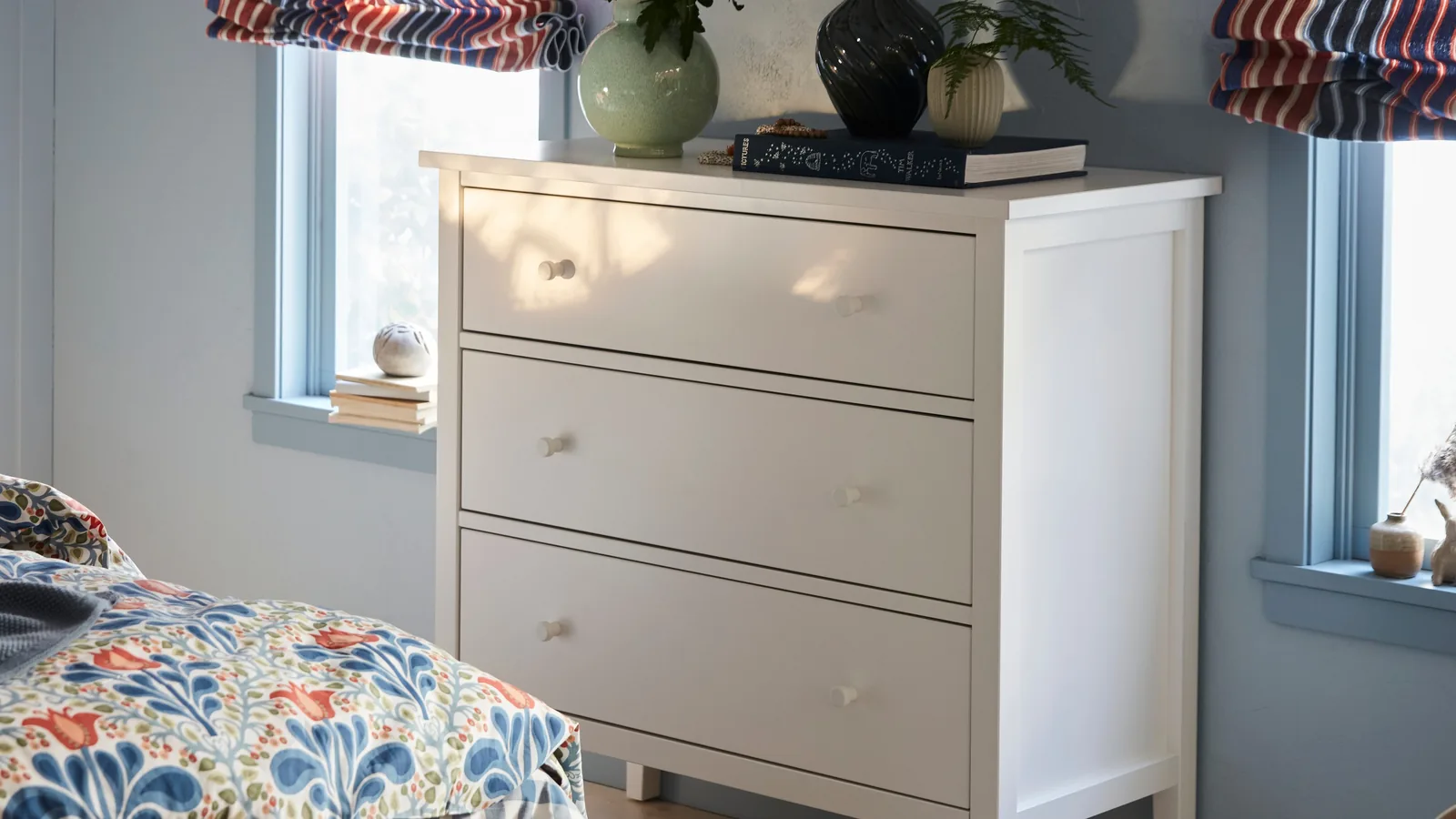 A white three-drawer chest of drawers topped with vases and books in a pale blue bedroom